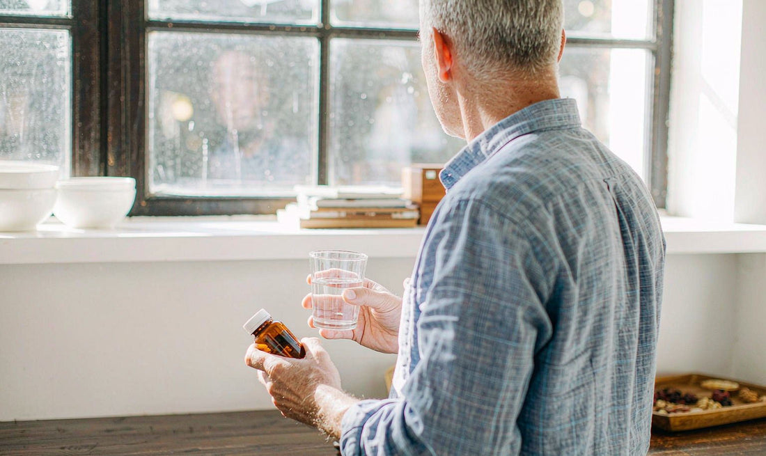 A man in his sixties seen from behind at a bright kitchen counter holding an amber supplement bottle and water