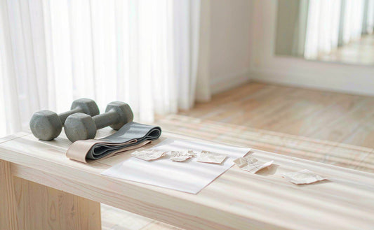 A pair of light dumbbells and a resistance band on a pale bench in a bright airy home gym corner