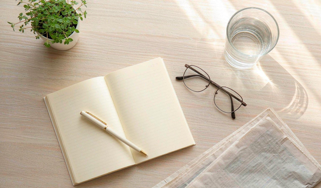 Overhead flat lay of an open weekly planner, reading glasses, water and a potted herb on pale oak