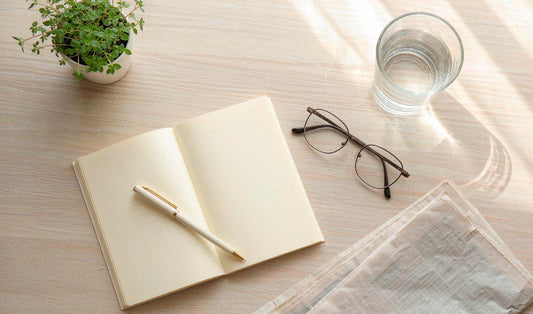Overhead flat lay of an open weekly planner, reading glasses, water and a potted herb on pale oak