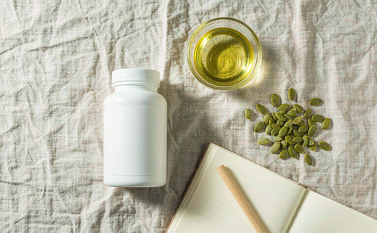 A flat lay of a blank supplement bottle, pumpkin seeds, oil and a notebook on pale linen