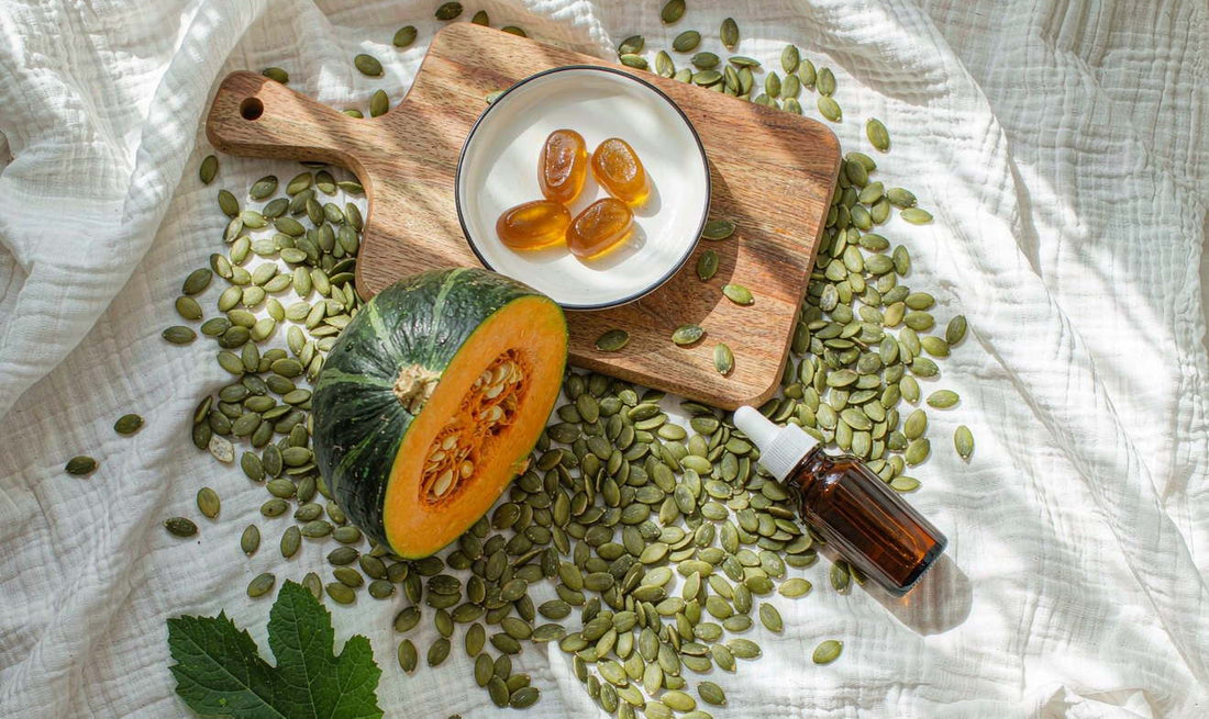 Flat lay of pumpkin seeds, a small amber bottle of seed oil and amber gummies beside half a fresh pumpkin