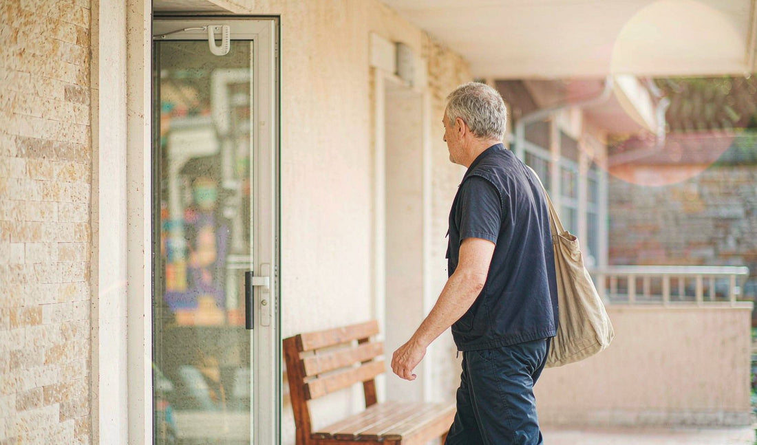 A man in his late fifties seen from behind in a navy volunteer vest, walking to a sunlit entrance