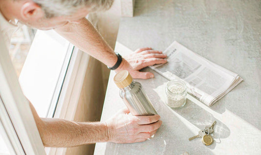 A man's hands setting a flask and a jar of seeds on a sunlit kitchen counter in the morning