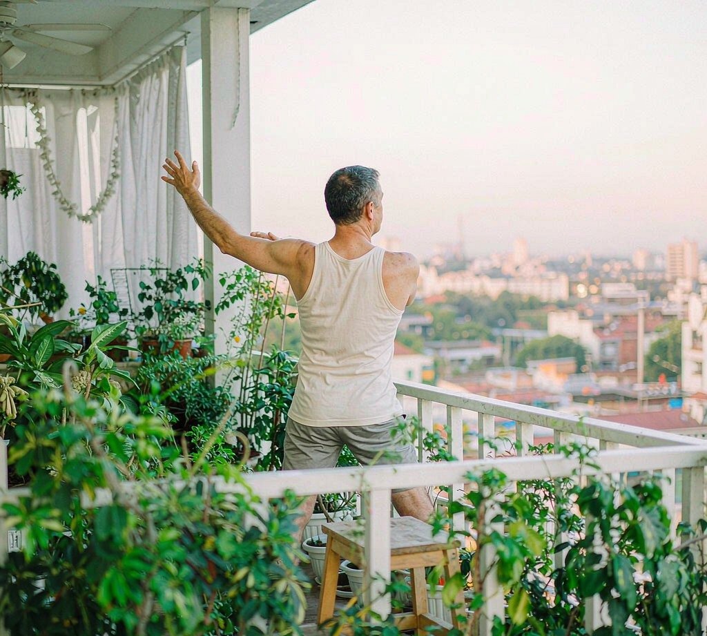 A man in his 50s stretching on a bright airy balcony at sunrise, seen from behind