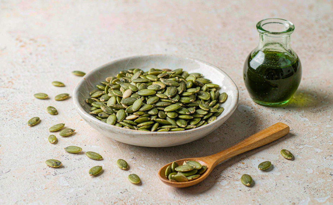A bowl of green pumpkin seeds with a wooden spoon and a small jug of dark oil in daylight