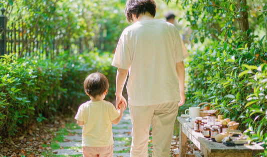 An adult hand holding a small child's hand from behind on a sunlit garden path with an amber jar